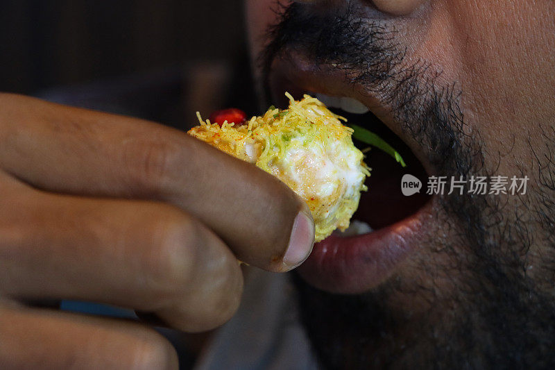 特写照片，一个认不出来的人满嘴吃着茶点，拿起golgappa puri(油炸水球)，空心酥脆的球体，里面有土豆、洋葱、石榴籽、辣味酸辣酱、Dahi papri chaat、酸奶和Sev